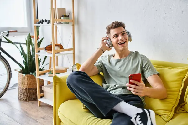 Encantador hombre con auriculares en sofá amarillo en casa desplazándose a las redes sociales y mirando a un lado - foto de stock