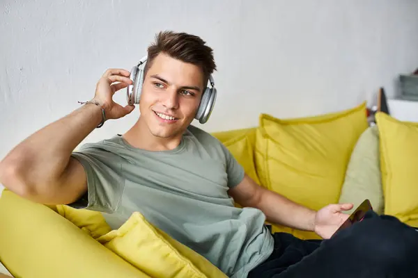 Charismatic man with headphones and smartphone in yellow couch at home looking to window — Stock Photo