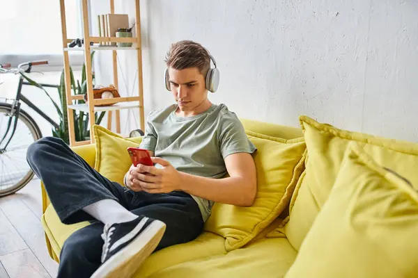 Joven estudiante en auriculares sentado en el sofá amarillo en casa y mensajes de texto en el teléfono inteligente - foto de stock