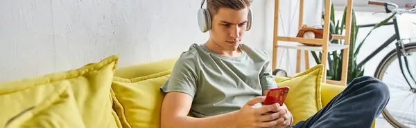Disparo horizontal de estudiante en auriculares sentados en el sofá amarillo en casa y mensajes de texto en el teléfono inteligente - foto de stock