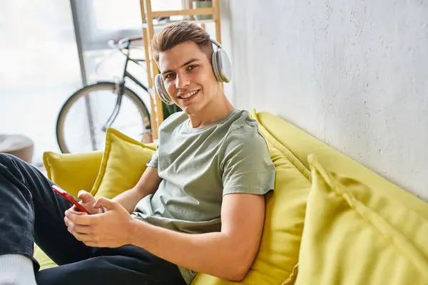 Hombre sonriente en auriculares en sofá amarillo en casa mensajes de texto en el teléfono inteligente y mirando a la cámara - foto de stock