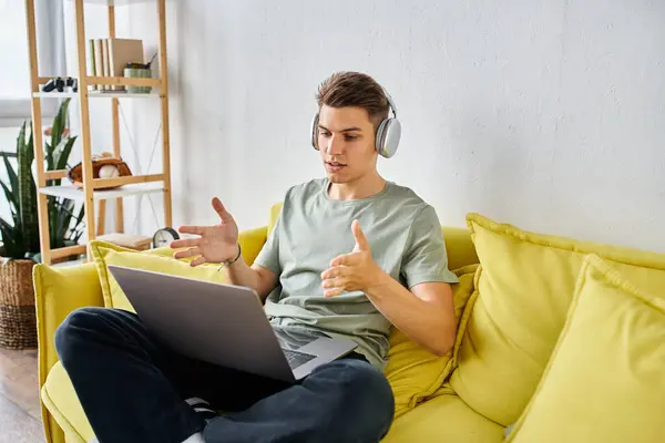 Estudiante seguro de sí mismo con auriculares en el sofá amarillo en casa hablando a la reunión en línea en el ordenador portátil - foto de stock