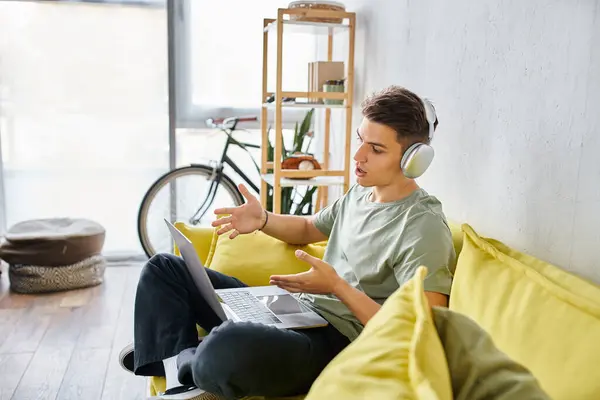 Perfil del tipo con auriculares en sofá amarillo en casa hablando de la reunión en línea en el ordenador portátil - foto de stock