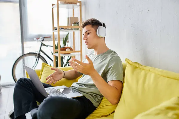 Estudiante enfocado con auriculares en el sofá amarillo en casa hablando a la reunión en línea en el ordenador portátil - foto de stock