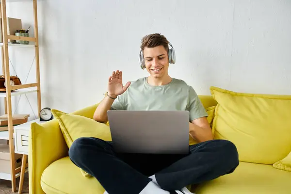 Cheerful student with headphones and laptop in yellow couch saying hello to online meeting — Stock Photo