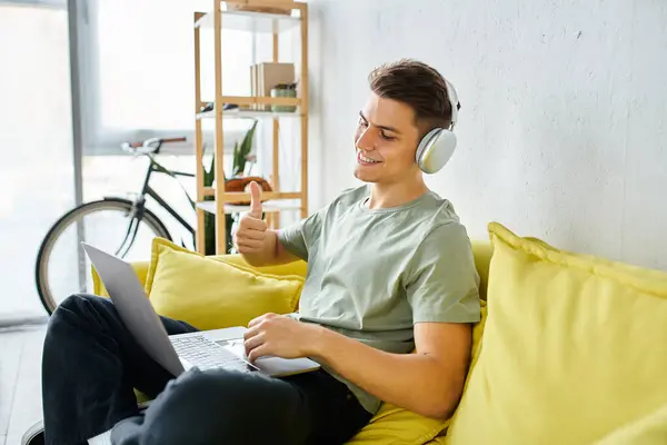 Усміхнений молодий чоловік з навушниками і ноутбуком в жовтому дивані любить онлайн-зустріч — стокове фото