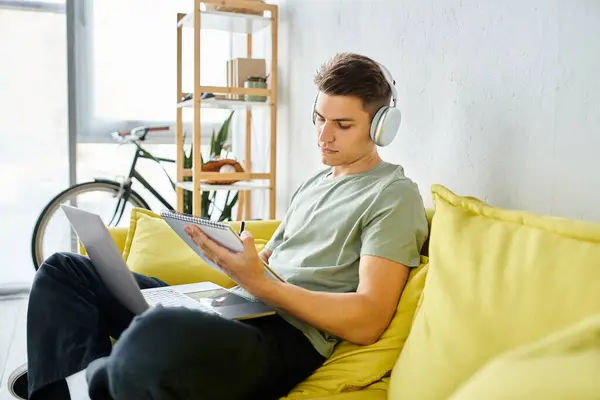 Encantador joven estudiante con auriculares y portátil en sofá amarillo estudiar y escribir en nota - foto de stock