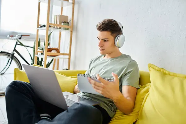 Estudiante seguro de sí mismo con auriculares en sofá amarillo estudiando en el ordenador portátil y leyendo en la nota - foto de stock