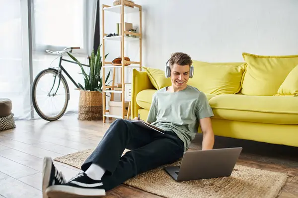 Lächelnder junger Mann mit Kopfhörern auf dem Boden neben gelbem Sofa, der in Laptop studiert und Notizen schreibt — Stockfoto