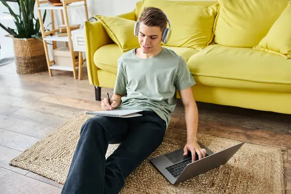 Student with headphones on floor near yellow couch studying in laptop and writing in note — Stock Photo