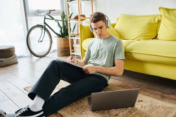 Сфокусированный студент с наушниками на полу рядом с желтым диваном, учится в ноутбуке и пишет заметки — стоковое фото