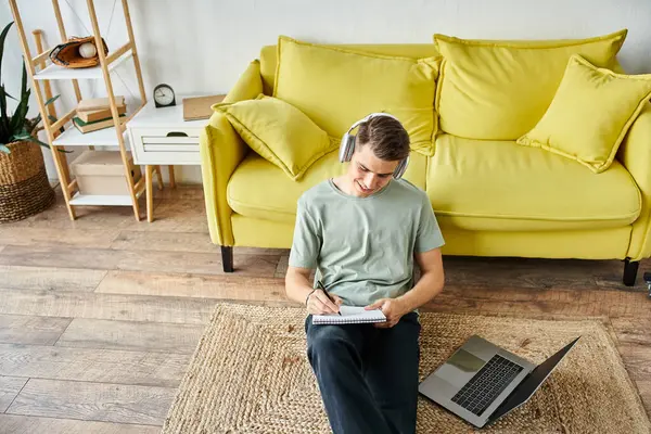 Lächelnder Kerl mit Kopfhörer und Laptop auf dem Boden in der Nähe der gelben Couch beim Lernen und Schreiben von Notizen — Stockfoto