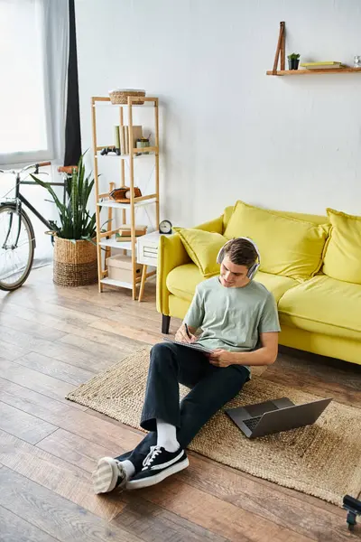 Cheerful young student with headphones and laptop on floor near yellow couch writing in note — Stock Photo