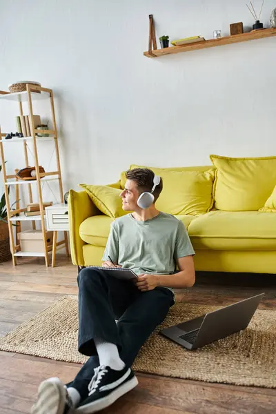 Mann mit Kopfhörer und Laptop auf dem Boden in der Nähe der gelben Couch schreibt Notizen und schaut zum Fenster — Stockfoto