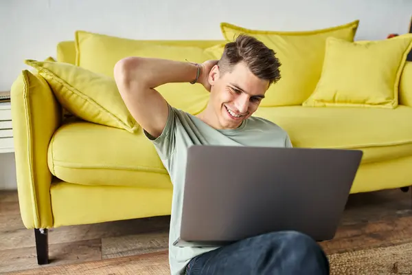 Confused student on floor near yellow couch at home studying in laptop and putting hand behind head — Stock Photo
