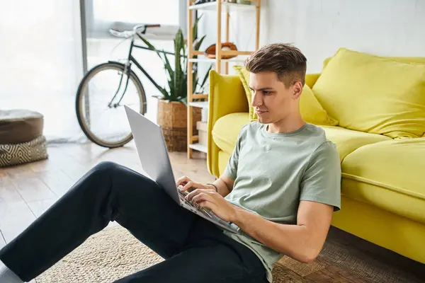 Atractivo estudiante de 20 años en el suelo cerca de sofá amarillo en el hogar en red en el ordenador portátil de lado - foto de stock