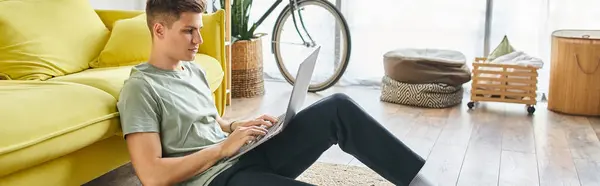 Banner of handsome man in 20s with brown hair on floor near yellow couch at home studying in laptop — Stock Photo