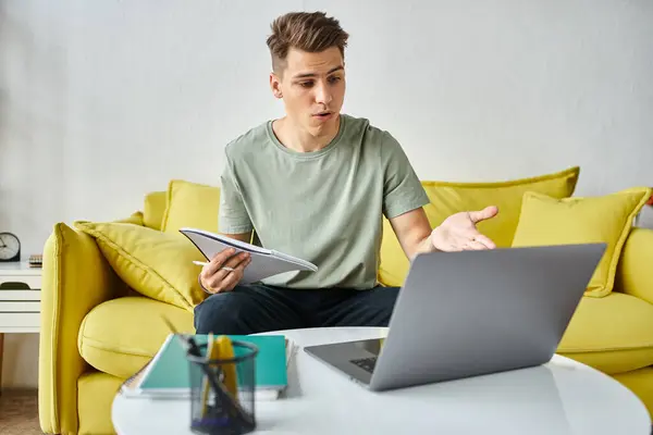 Заплутаний молодий студент в жовтому дивані вдома виконує курсову роботу в нотатках і ноутбук на кавовому столі — стокове фото