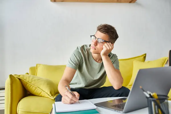 Bored charming man with glasses in yellow couch at home writing in notes and looking to window — Stock Photo