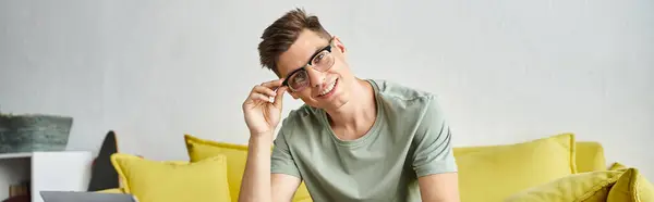 Banner of smiling man in 20s with vision glasses on yellow couch putting pen down on coffee table — Stock Photo