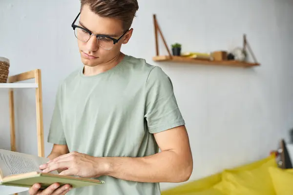 Attractive student in his 20s with brown hair and vision glasses in living room reading book — Stock Photo