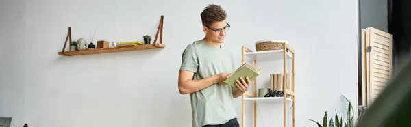 Horizontal shot of young man with brown hair and vision glasses in living room reading book — Stock Photo