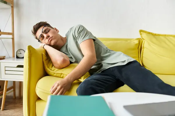 Уставший молодой студент с каштановыми волосами и очками зрения спит на желтом диване в гостиной — стоковое фото