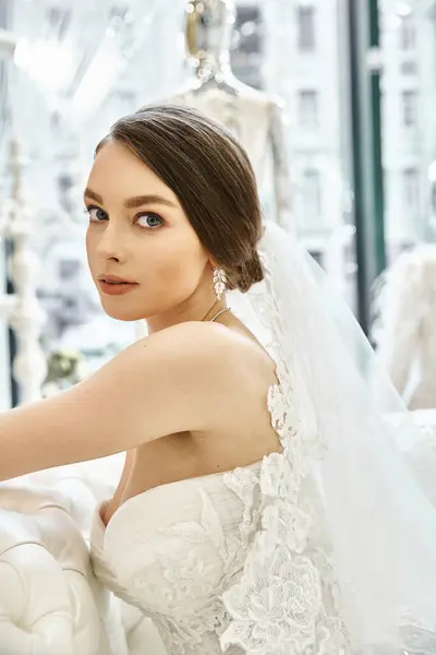 A young brunette bride in a flowing white wedding dress, seated gracefully on a luxurious couch in a bridal salon. — Stock Photo