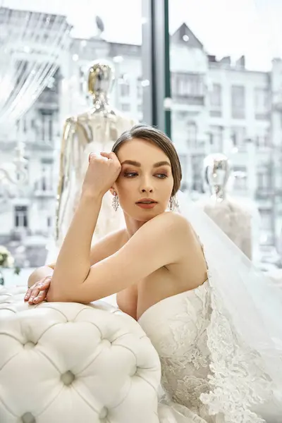 A young brunette bride in a wedding dress gracefully sits on a elegant couch in a bridal salon. — Stock Photo