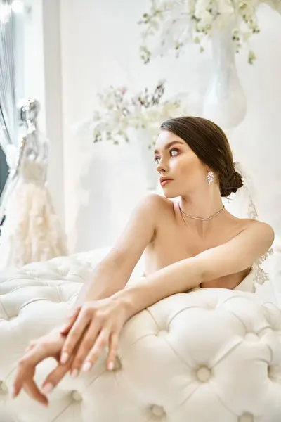 Eine junge brünette Braut in ihrem Brautkleid sitzt anmutig auf einem weißen Bett, umgeben von einer Aura der Ruhe und Schönheit. — Stockfoto