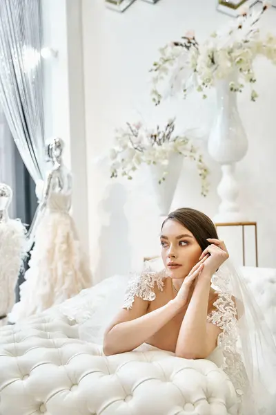Une jeune mariée brune dans une robe de mariée posée paisiblement sur un lit blanc immaculé. — Photo de stock