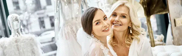 Eine junge brünette Braut im Brautkleid und ihre blonde Mutter mittleren Alters stehen nebeneinander in einem Brautsalon. — Stockfoto