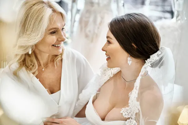 A young brunette bride in a wedding dress is standing next to her middle-aged blonde mother in a bridal salon. — Stock Photo