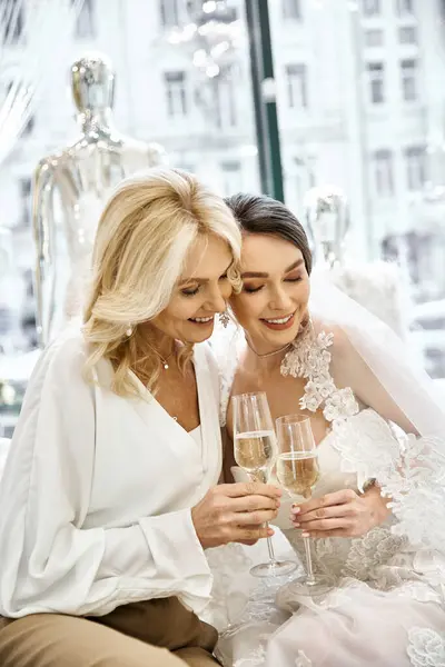 Молодая невеста в свадебном платье и ее мать сидят вместе, держа бокалы вина в свадебном салоне. — стоковое фото