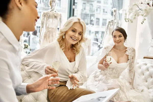 A young brunette bride in her wedding dress sits next to her bridesmaid and middle-aged blonde mother on a cozy couch in a bridal salon. — Stock Photo