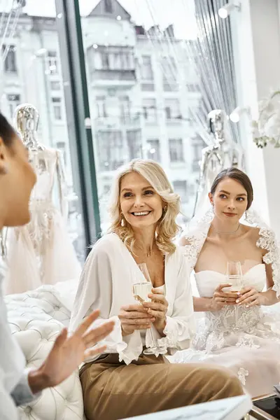 Una joven novia con un vestido de novia, su dama de honor, y una madre de mediana edad sentada de cerca en un salón de novias. - foto de stock