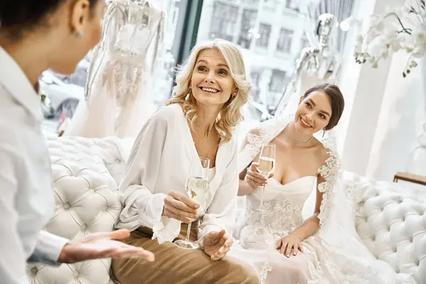 Молода брюнетка наречена у весільній сукні, її невільниця та блондинка середнього віку у весільному салоні, сидить разом. — стокове фото