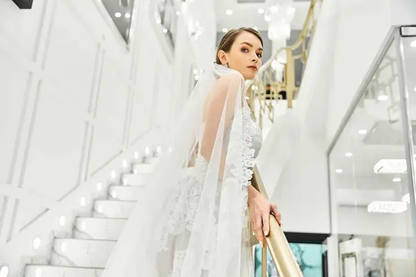 A young brunette bride, dressed in a wedding gown, standing gracefully on a staircase in a bridal salon. — Stock Photo