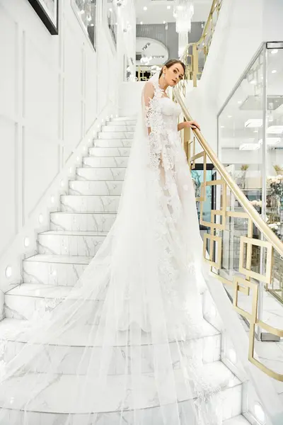 A young brunette bride in a wedding dress stands on a staircase in a bridal salon. — Stock Photo