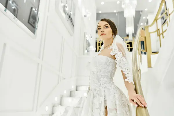A young brunette bride in a wedding dress stands on a staircase in a bridal salon. — Stock Photo
