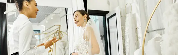 Une jeune mariée brune en robe de mariée et son assistante shopping debout devant un miroir dans un salon de mariée. — Photo de stock