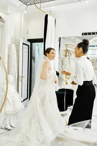 Молодая невеста в белом платье и ассистент стоят вместе, глядя в зеркало в свадебном салоне. — стоковое фото