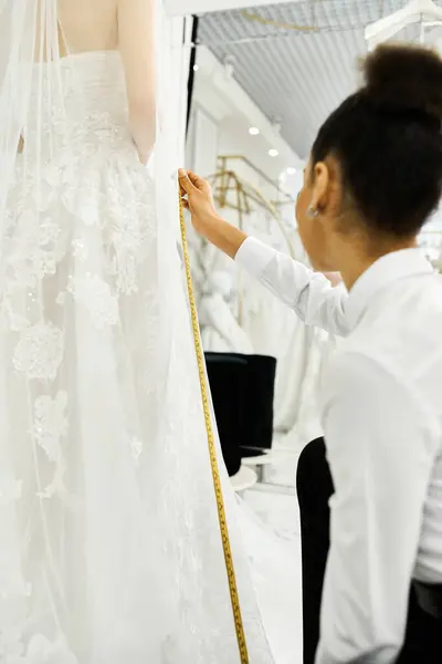 Молодая невеста измеряет свадебное платье на манекене с помощью афроамериканского торгового ассистента в свадебном салоне. — стоковое фото