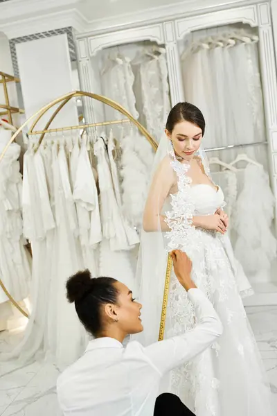 Uma jovem noiva morena em um vestido de noiva está sendo medida por um assistente de compras afro-americano em um salão de noivas. — Fotografia de Stock