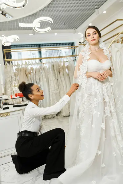 Eine junge Braut im Brautkleid kniet neben ihrer afroamerikanischen Shopping-Assistentin in einem Brautsalon. — Stockfoto