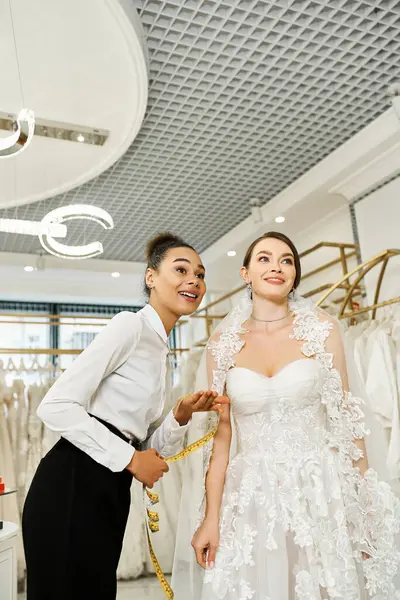 Young brunette bride in a white wedding dress standing next to an African American woman in a black dress in a bridal salon. — Stock Photo