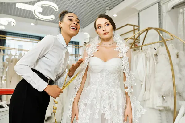 Eine junge brünette Braut im Brautkleid steht neben einer afroamerikanischen Shopping-Assistentin in einem Brautsalon. — Stockfoto