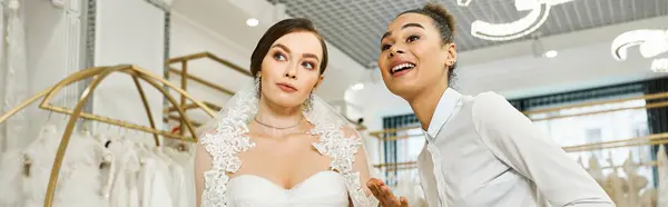 Eine junge brünette Braut im Brautkleid wird von einer afroamerikanischen Shopping-Assistentin in einem Brautsalon begleitet. — Stockfoto