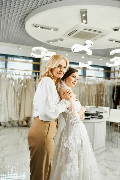Eine junge brünette Braut im Brautkleid steht mit ihrer Mutter mittleren Alters in einem Brautsalon und bewundert Kleider. — Stockfoto