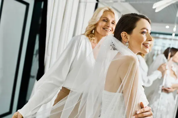 Una joven novia en un vestido de novia está de pie con su madre de mediana edad en un salón de novias, rodeado por un grupo de mujeres. - foto de stock